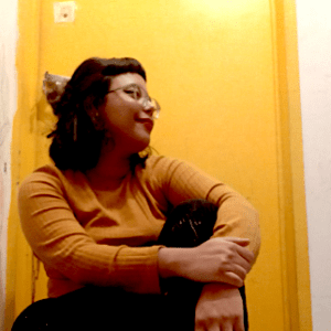 profile-picture mujer, viendo a la derecha con un sueter amarillo, sentada en el suelo con las piernas cruzadas. Imagen de perfil